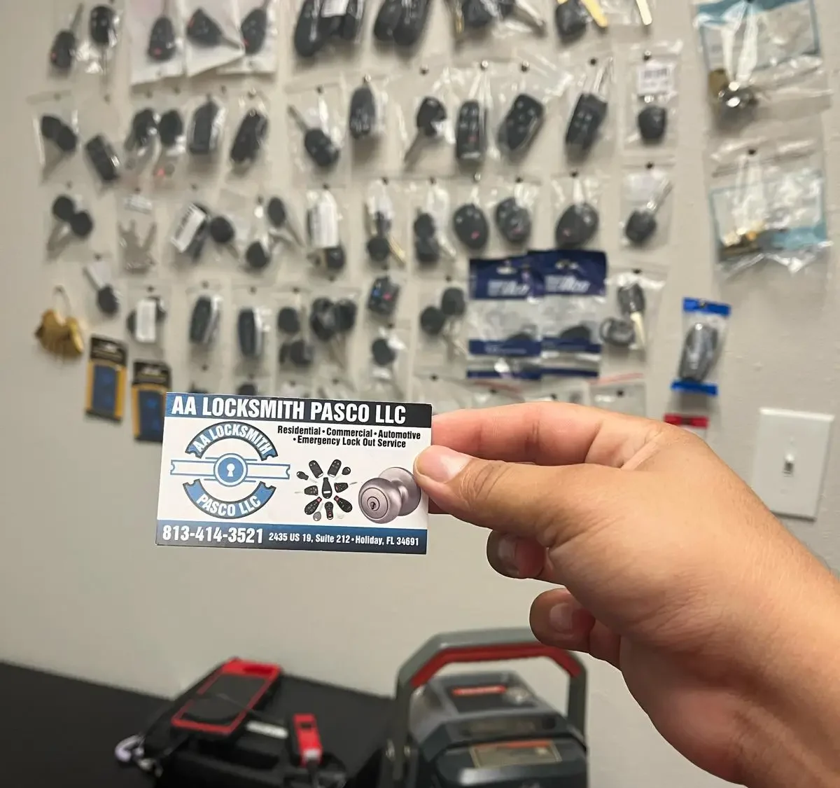 A person holds a business card for a locksmith service in front of a wall filled with various tools.